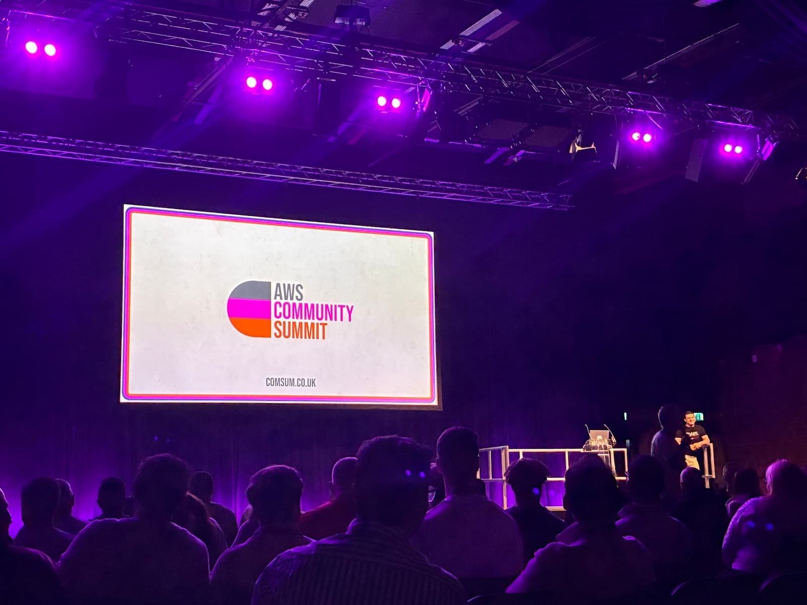 The AWS Community Summit logo on the main stage screen at Victoria Warehouse, Manchester, with the audience in the foreground